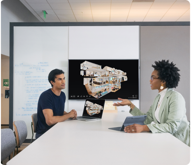 Two people sitting on opposite sides of a conference table, showing each other their laptop screens, with a Matterport dollhouse view in the background
